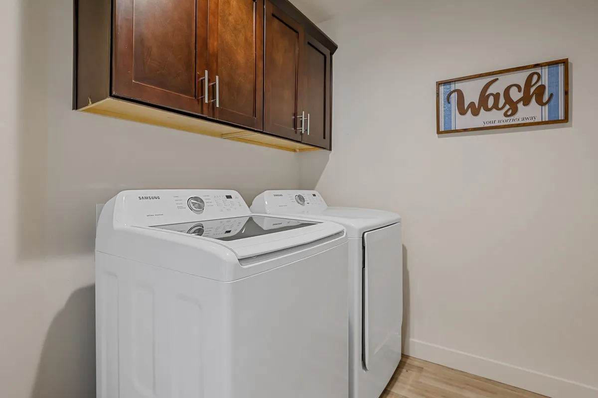 A well-lit laundry room features brand-new washing machines beneath wooden cabinets, with a decorative sign adding a homey touch. The space is tidy and organized, emphasizing cleanliness and ease of use.