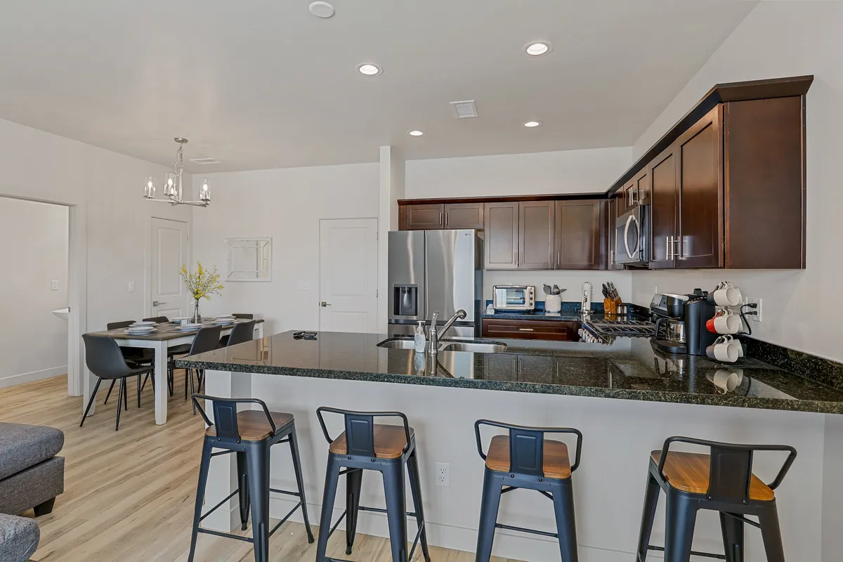 A contemporary, warm kitchen and dining area featuring a central kitchen island, sleek stainless steel appliances, and a spacious dining table for six. The space is brightly lit and arranged for both cooking and entertaining.
