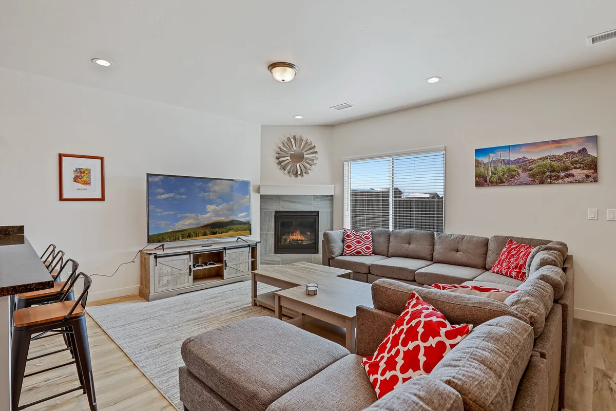 A spacious living room scene featuring a large sectional sofa positioned in front of a warm fireplace. The area is designed for comfort and gathering, with a massive TV mounted on the wall and stylish decorative wall art completing the inviting ambiance. The layout encourages both entertainment and relaxation, perfect for family movie nights or cozy evenings by the fire.