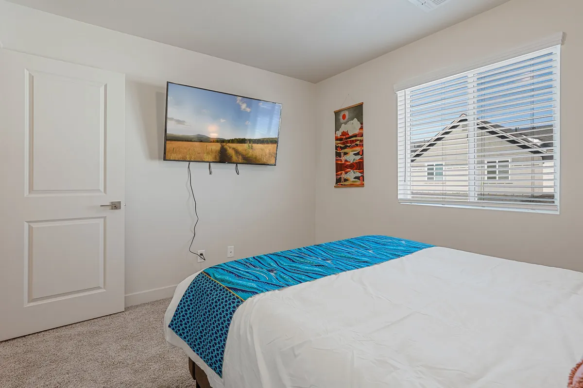 A primary bedroom featuring a king-sized bed with blue and white bedding, a wall-mounted Smart TV, a window with blinds allowing natural light, and decorative wall art above the bed. The room exudes a cozy and modern vibe, with plush bedding and blackout curtains suggesting restful comfort.