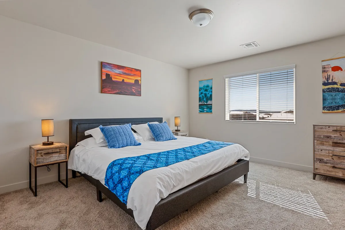 A serene bedroom with a queen-sized bed dressed in blue and white bedding, complemented by tasteful wall art and bedside lamps. A window with blinds lets in soft morning light, creating a calm and inviting atmosphere ideal for relaxation and reading.