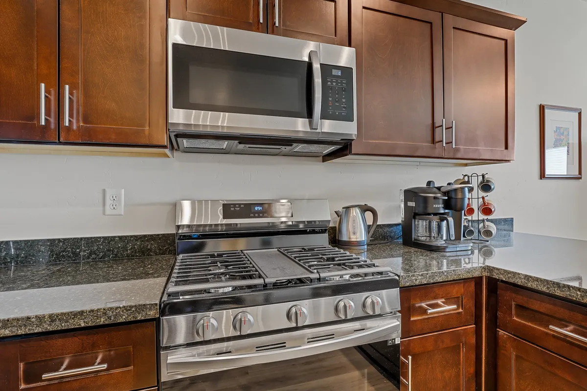 A close-up view of a stainless steel stove with granite countertops, a prominently placed coffee machine (likely a Keurig), and bright, cozy kitchen lighting. The scene is set for peaceful coffee moments and meal creation.