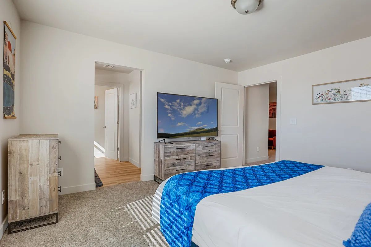 A cozy bedroom setup featuring a bed with a blue patterned blanket, a Smart TV, and a wooden dresser. The window brings in natural light, making the room inviting and comfortable for unwinding after work or catching up on shows.