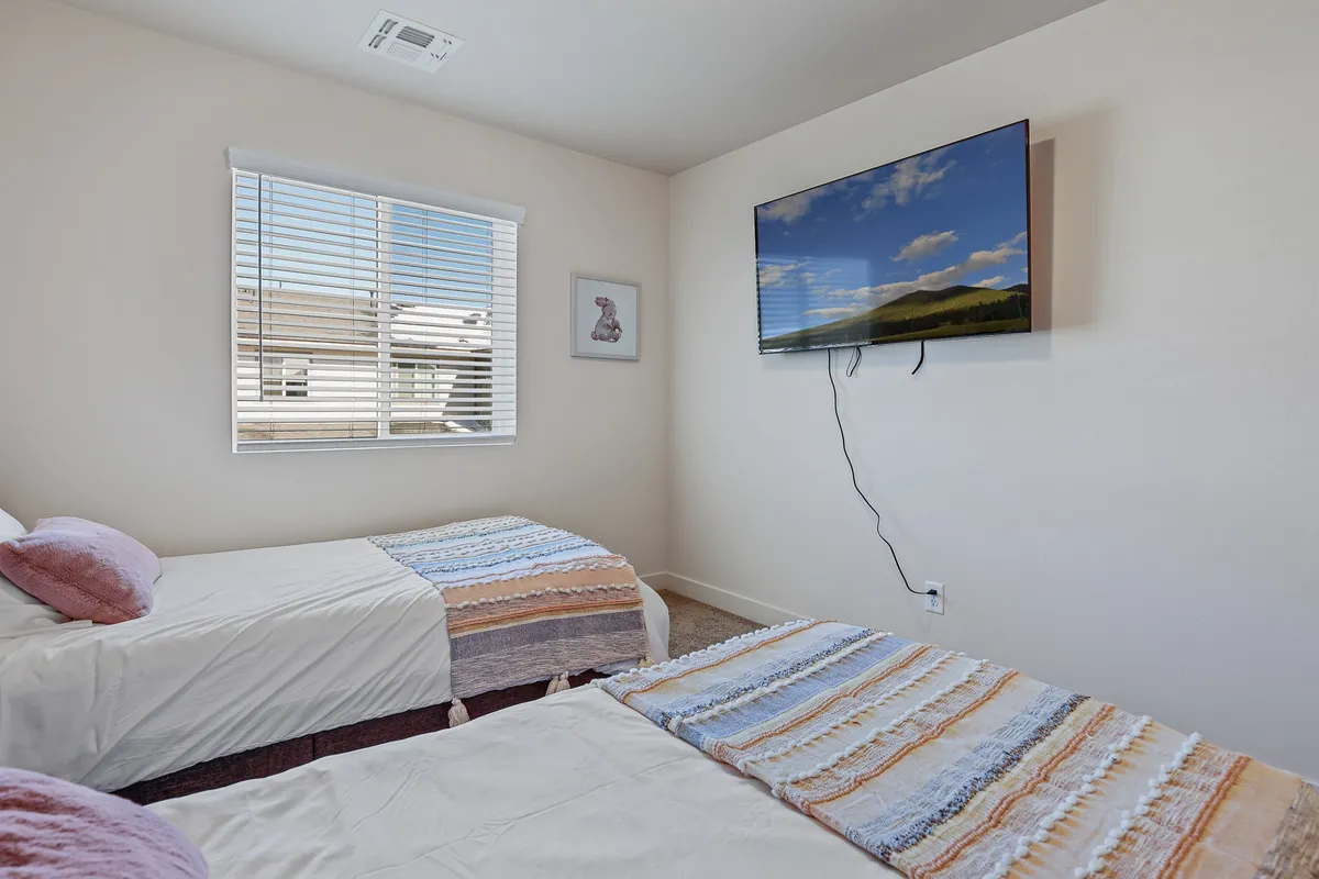 A cozy bedroom setup featuring twin beds, a Smart TV, and a window with blinds. The room is bathed in soft light, offering a comfortable space for relaxing, watching TV, and enjoying a quiet moment.