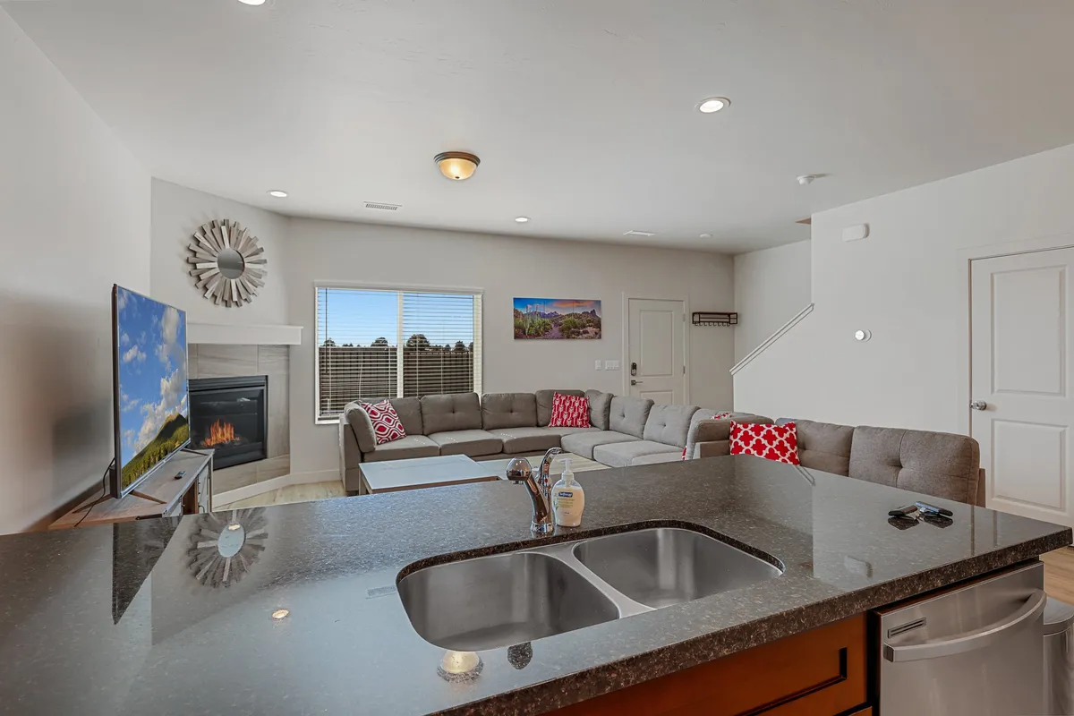 A bright, comfortable living area with a sprawling sectional sofa, a large TV, a fireplace, and a decorative mirror. The open-plan arrangement also shows the kitchen island in the background, conveying a warm, homey atmosphere ideal for group entertainment and relaxation.